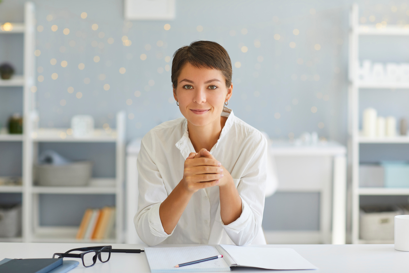 Confident Lady Looking at Camera Sitting in Her Home Office at Desk with Open Notebook